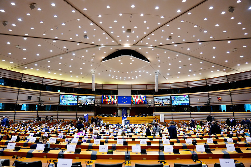 Blick in den modern gestalteten Plenarsaal des Europäischen Parlaments in Brüssel mit zahlreichen Sitzreihen, Monitoren, Namensschildern und EU- sowie Mitgliedstaatenflaggen vor dem Rednerpult.