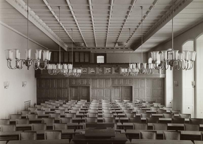 Schwarz-weiß-Fotografie eines Sitzungssaals mit Holzvertäfelung, Reihen schlichter Stühle und mehreren Kronleuchtern – Nachkriegszeit