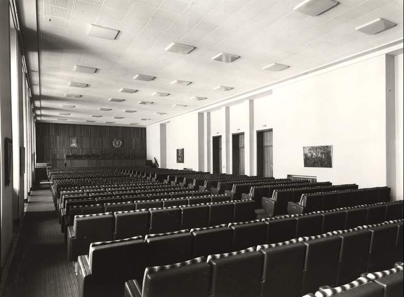 Saal mit Reihen gepolsterter Stühle und flacher Decke mit quadratischen Leuchten – funktional gestalteter Sitzungssaal der 1970er Jahre