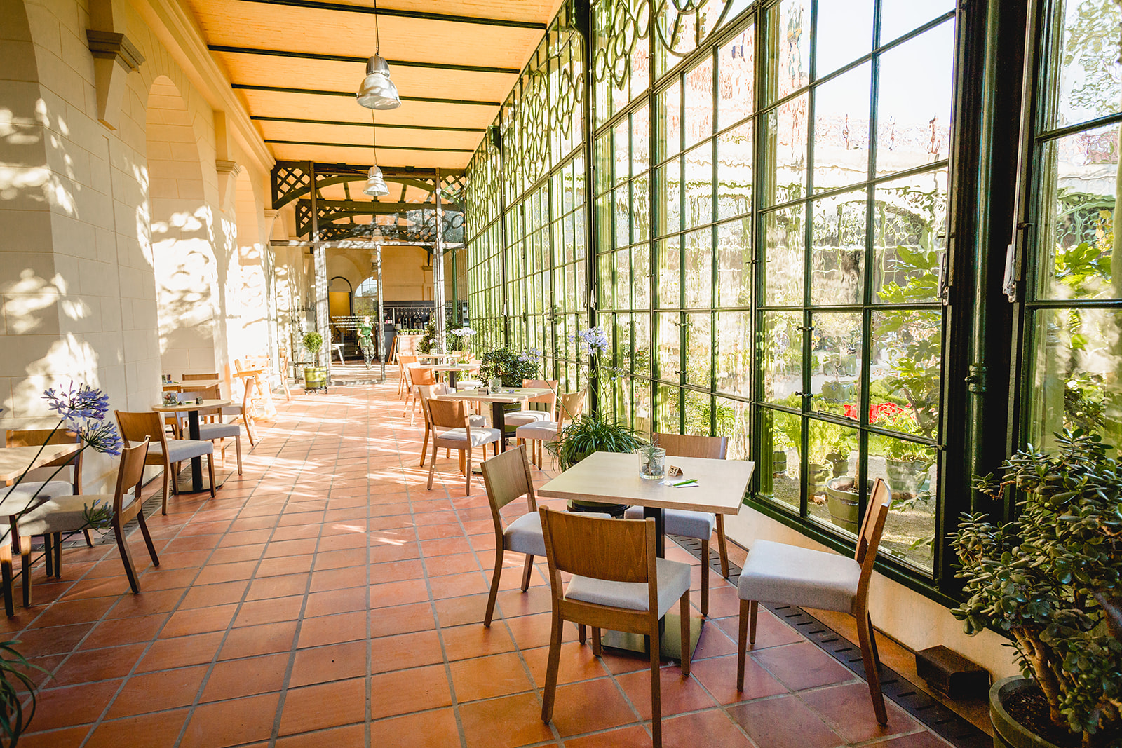 Ein heller Wintergarten, die Orangerie des Schlosses, mit großen Fenstern, Terrakottafliesen und mehreren kleinen, modern gedeckten Tischen mit Holzstühlen und Pflanzen.
