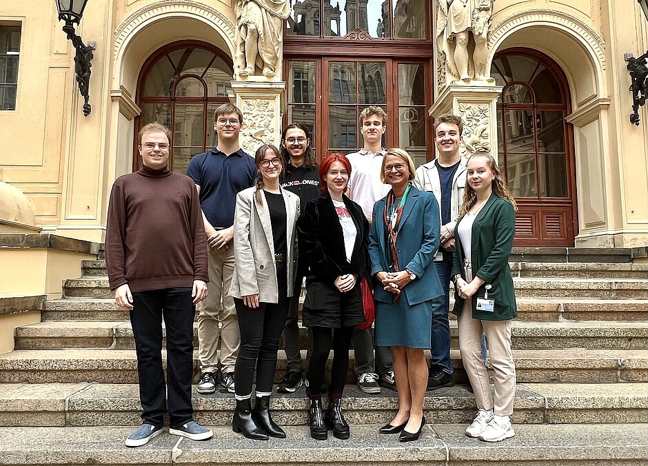 Acht junge Erwachsene und eine Frau in einem blauen Kostüm, stehen auf einer Treppe vor dem Eingang des Schweriner Schlosses und posieren für ein Gruppenfoto.
