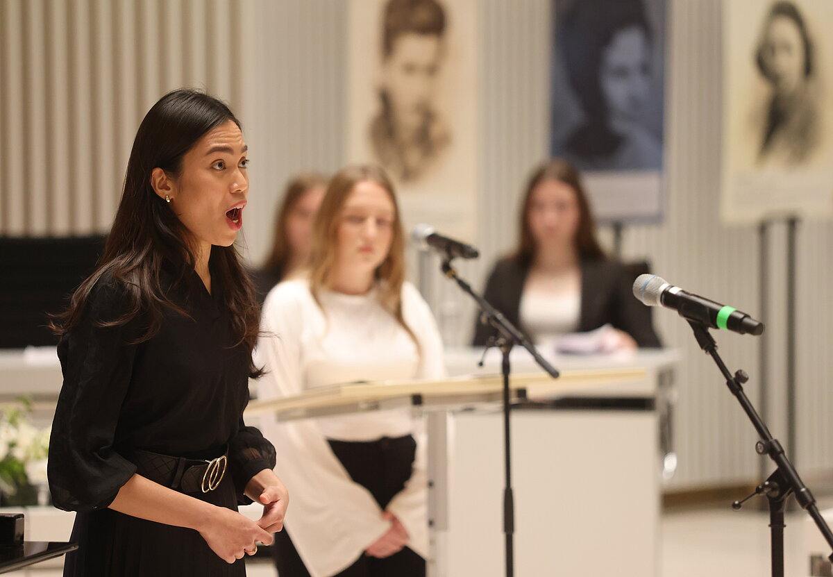 Portraitaufnahme einer Frau, die singt; im Hintergrund zwei Schülerinnen am Rednerpult des Plenarsaals