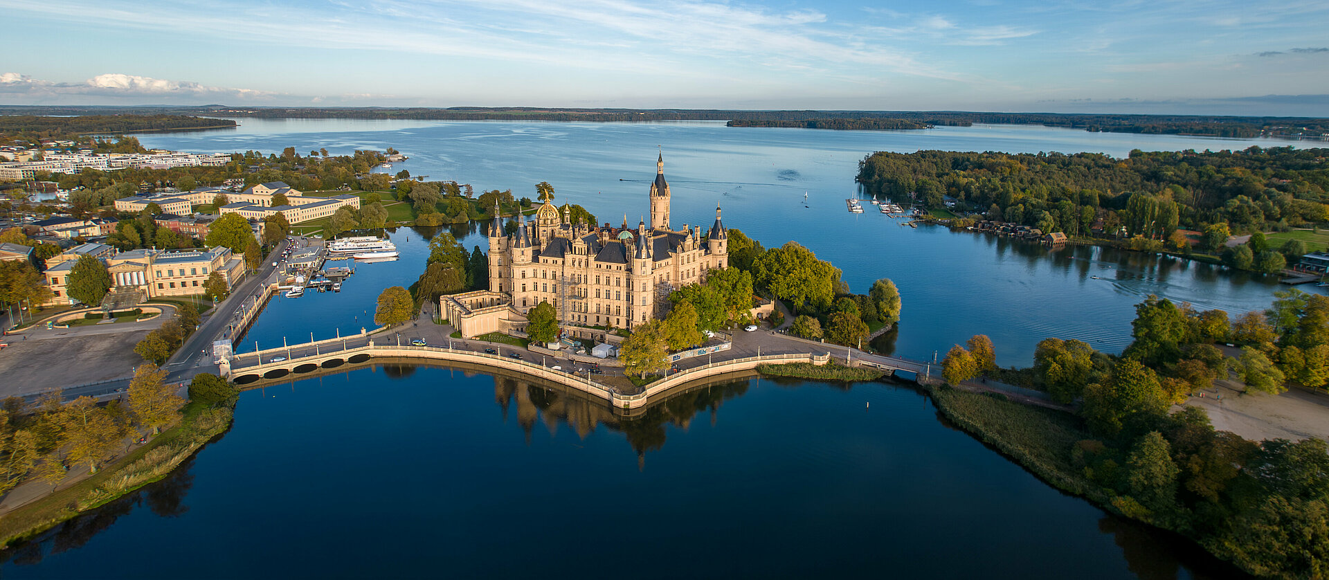 Luftaufnahme des Schweriner Schlosses, das von Wasser umgeben auf einer Insel im Schweriner See liegt. Umgeben von Bäumen, Brücken und angrenzenden historischen Gebäuden, erstreckt sich im Hintergrund die Stadt Schwerin.