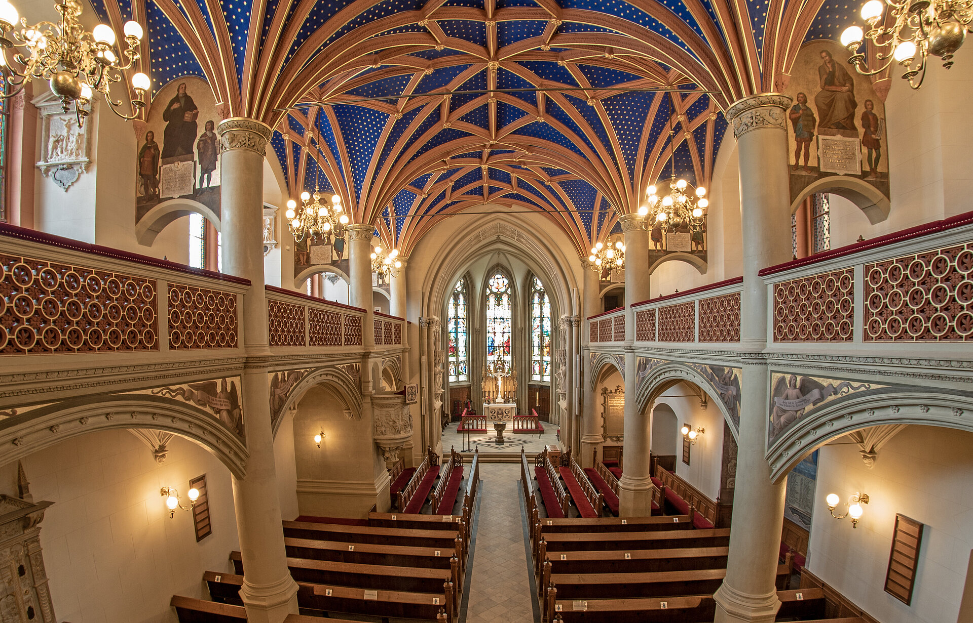 Blick in das Kirchenschiff der Schlosskirche von einer Empore aus mit blauen Gewölbedecken, kunstvollen Leuchtern, Wandmalereien und Holzgestühl.