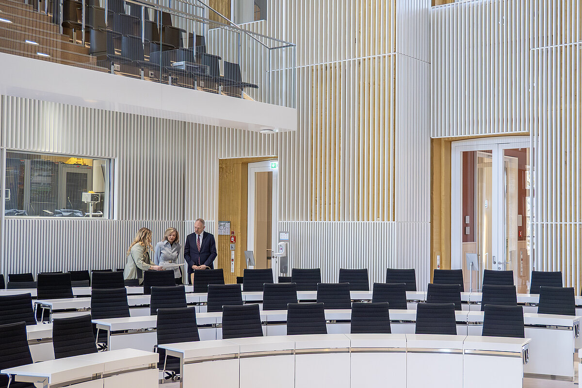 Drei Personen besichtigen einen hellen Plenarsaal, es sind leere Stuhlreihen der Abgeordneten zu sehen