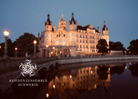 Abendliche Ansicht des beleuchteten Schweriner Schlosses mit Spiegelung im Schlossgraben und historischer Brücke im Vordergrund.