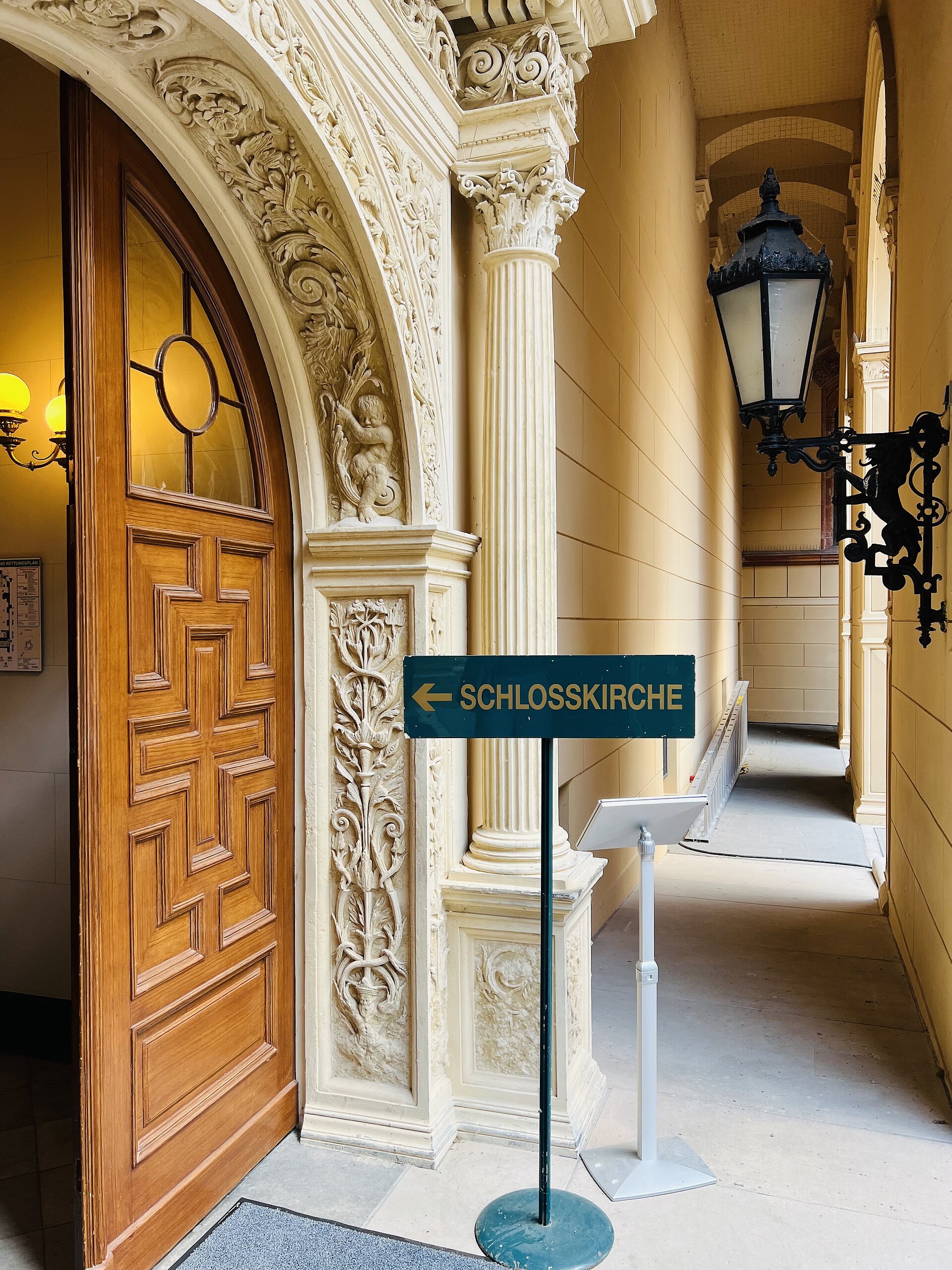 Eingang zur Schlosskirche mit geöffneter hölzerner Tür, reich verziertem Steinportal und einem Schild mit der Aufschrift „Schlosskirche“ auf einem Pfeilständer.