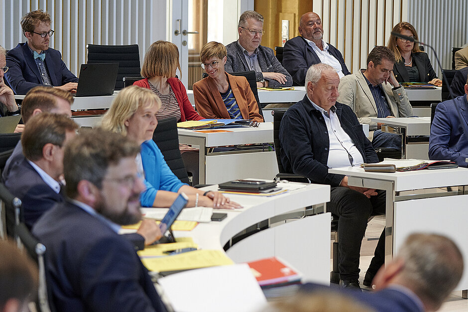 Abgeordnete sitzen in einem modernen Plenarsaal an geschwungenen Tischen und verfolgen konzentriert eine Sitzungmern während einer Sitzung