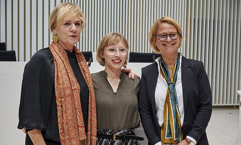 Drei Frauen stehen im Plenarsaal des Landtages und schauen zufrieden ins Publikum