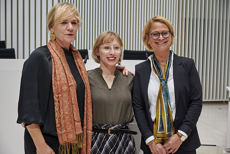 Drei Frauen stehen im Plenarsaal des Landtages und schauen zufrieden ins Publikum