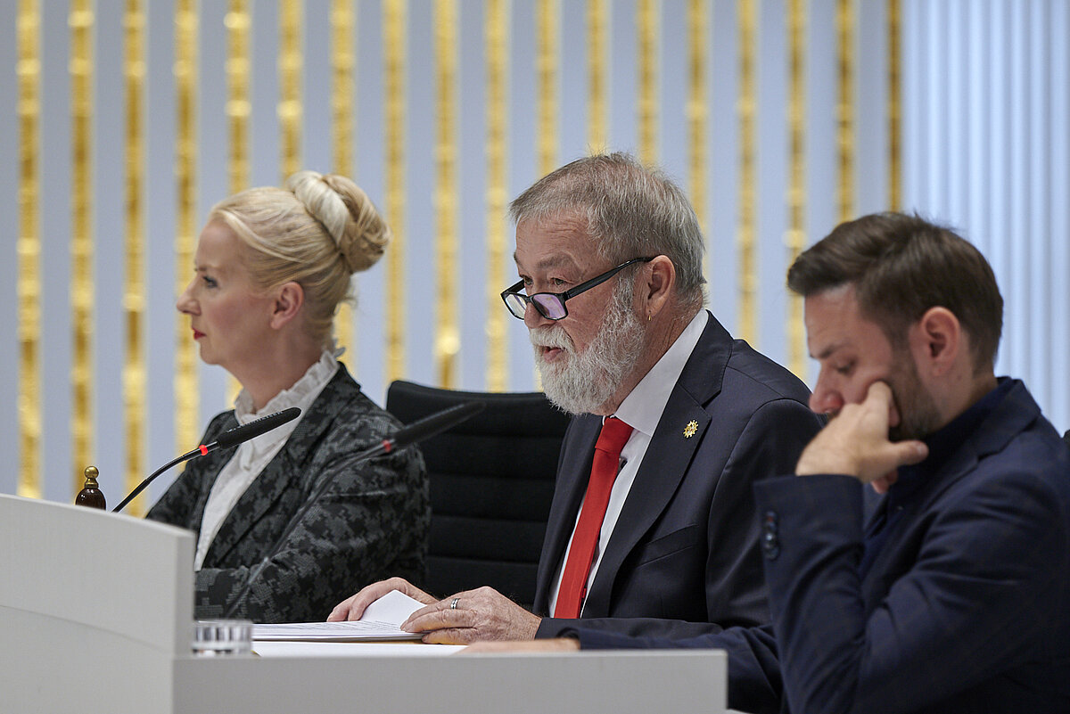Ein Mann mit Bart sitzt am Pult und spricht ins Mikrofon, neben ihm sitzen eine blonde Frau und ein jüngerer Mann