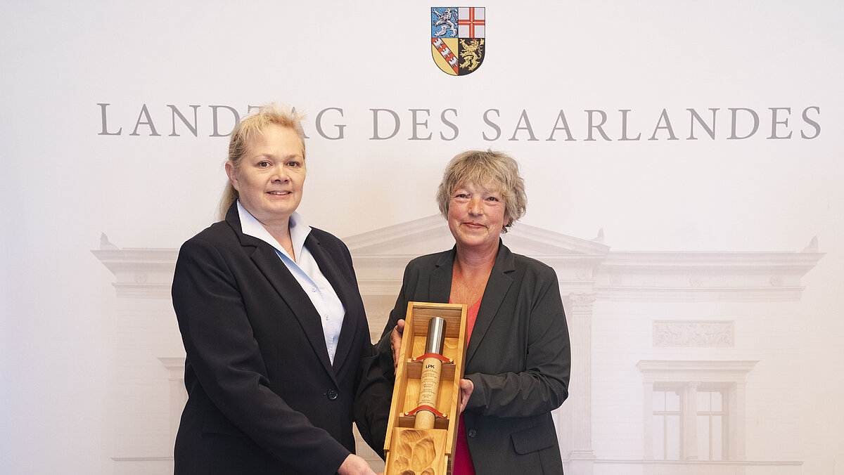 Zwei Frauen in Anzügen halten gemeinsam eine lange Holzbox vor einer Wand mit der Aufschrift 'Landtag des Saarlandes' und einem Wappen
