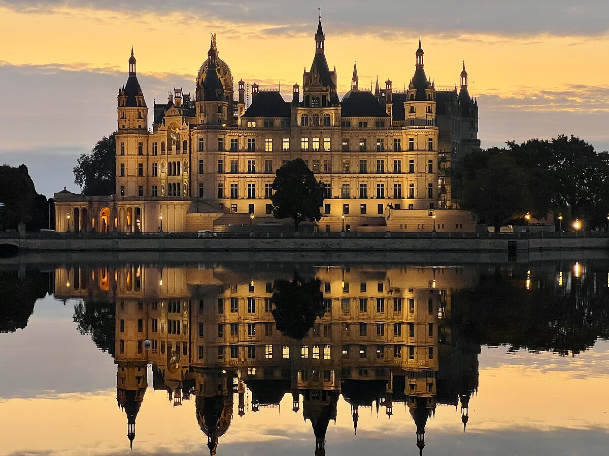 Abendliche Ansicht des angestrahlten Schweriner Schlosses mit beeindruckender Spiegelung im ruhigen Wasser des umgebenden Sees vor einem goldfarbenen Himmel bei Sonnenuntergang.