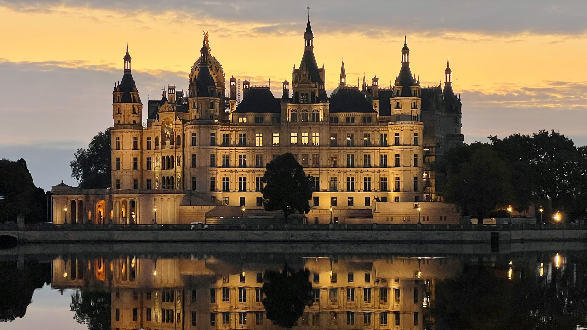 Abendliche Ansicht des angestrahlten Schweriner Schlosses mit beeindruckender Spiegelung im ruhigen Wasser des umgebenden Sees vor einem goldfarbenen Himmel bei Sonnenuntergang.