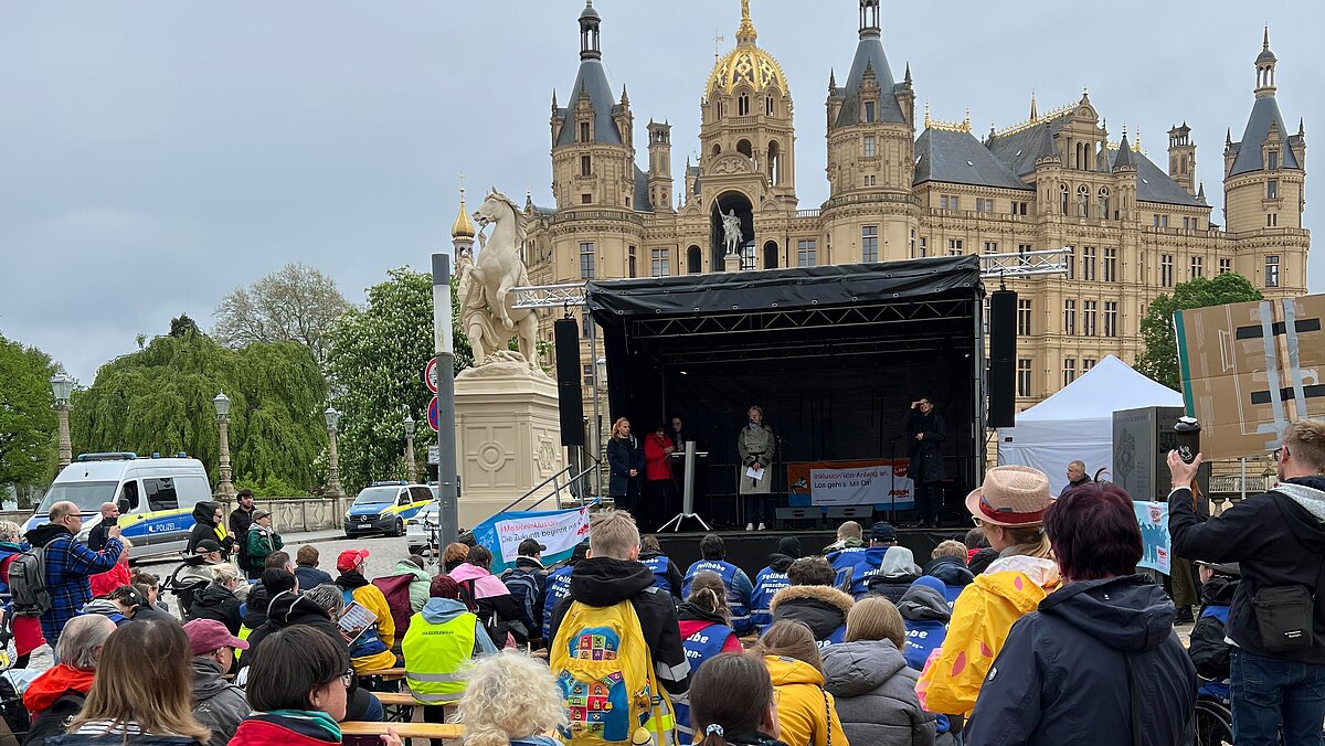 Menschen sitzen auf Bänken vor einer Bühne mit Rednern vor einem historischen Schlossgebäude bei bewölktem Himmel