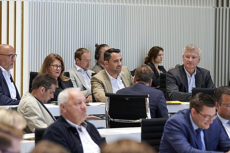 Abgeordnete verschiedener Fraktionen sitzen im Plenarsaal an weißen Tischen und verfolgen aufmerksam die laufende Debatte