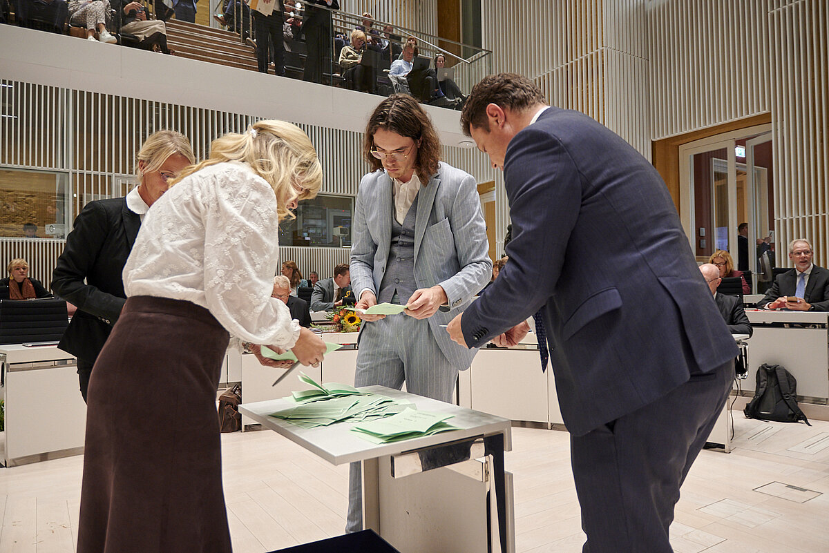 Mehrere Personen sichten Wahlzettel, eine Urne ist zu sehen.