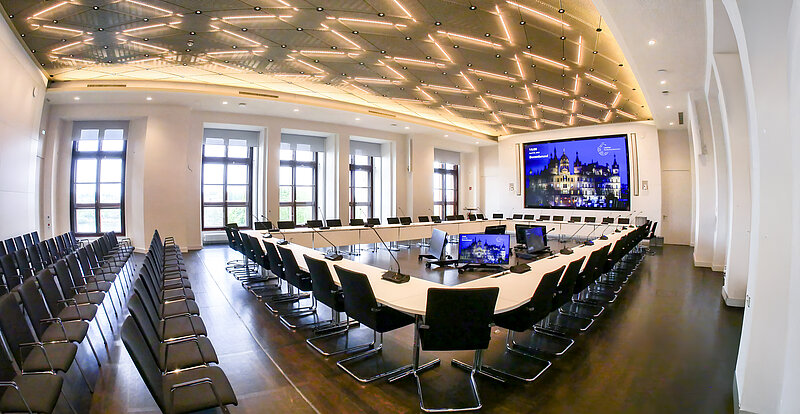 Moderner, heller Konferenzraum im Demmler-Saal des Landtags Mecklenburg-Vorpommern mit u-förmig angeordneten Tischen, Mikrofonen und Monitoren, umgeben von Stuhlreihen und großen Fenstern. Eine große Leinwand zeigt das Schweriner Schloss.