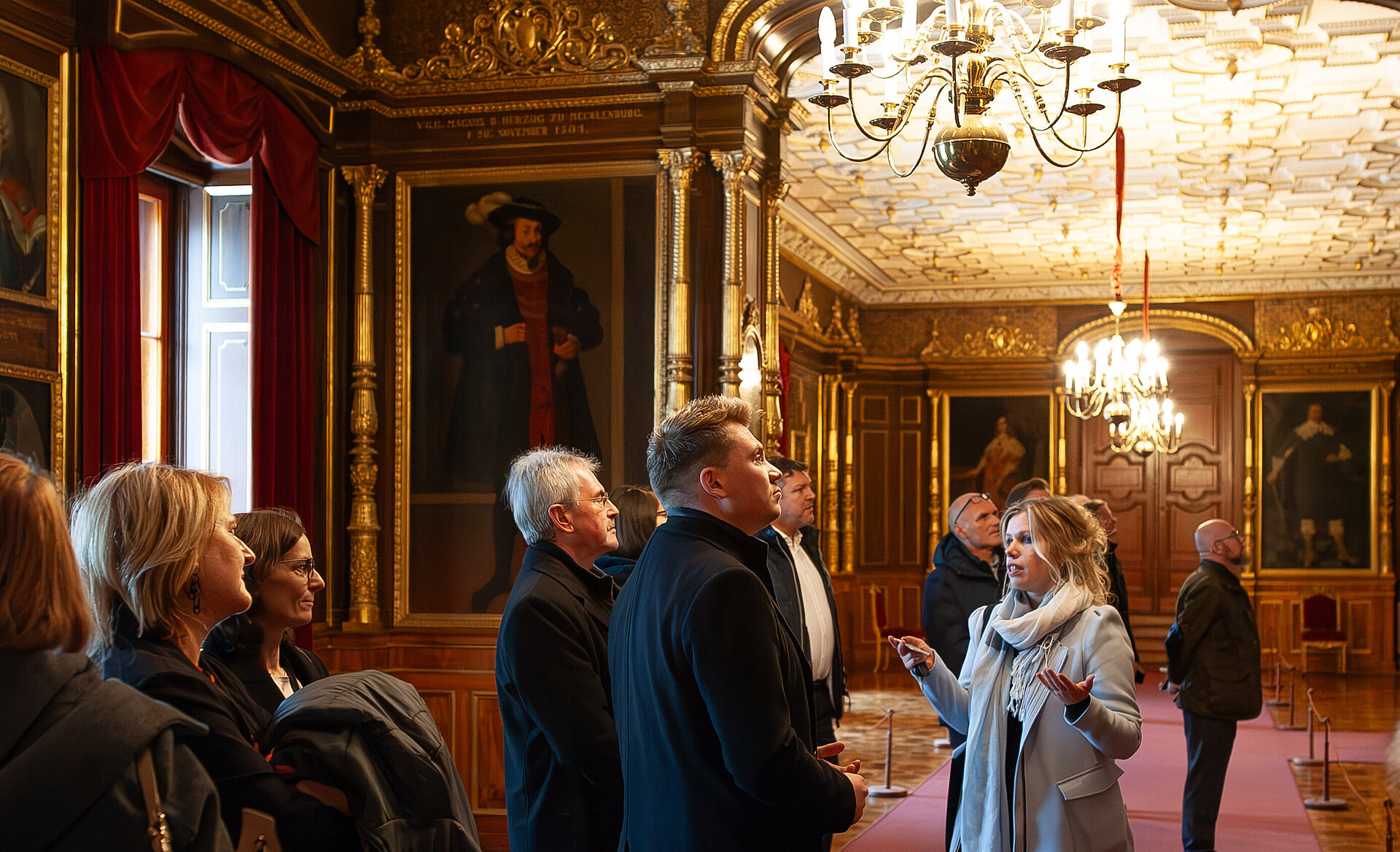 Eine Gruppe von Besucherinnen und Besuchern wird von einer Führerin durch einen prunkvoll ausgestatteten Raum mit goldenen Verzierungen, großen Porträts und Kronleuchtern geführt.