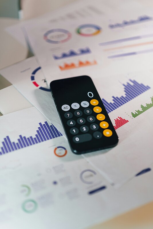 Papiere mit Statistiken und ein Smartphone mit Taschenrechner-App auf einem Tisch.