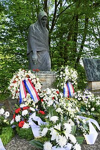 Mahnmal "Die Mutter" - Bronzefigur einer knienden Frau mit Kopftuch auf Steinsockel umgeben von Kränzen und Blumen in einem Wald