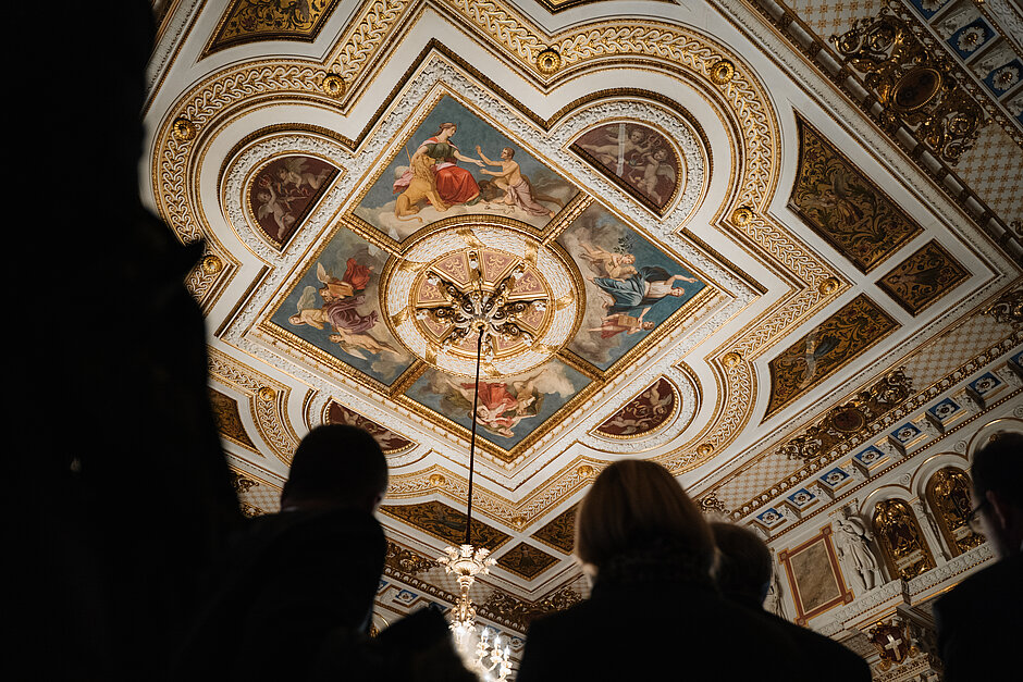 Blick einer Besuchergruppe auf eine reich verzierte Decke mit farbenfrohen Fresken und goldenen Ornamenten in einem historischen Schlossraum.