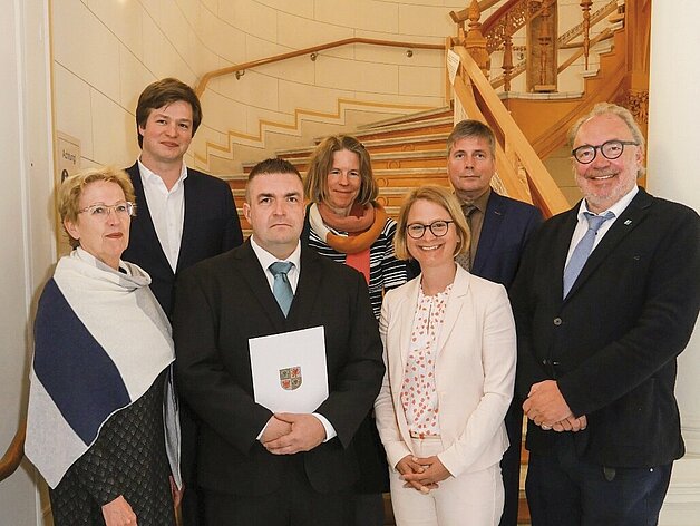 Gruppenfoto von acht Personen in formeller Kleidung vor einer geschwungenen Treppe im Landtag Mecklenburg-Vorpommern; der Datenschutzbeauftragte in der Mitte hält eine Mappe mit dem Landeswappen“