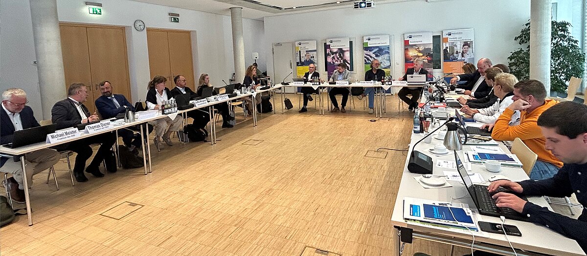 Mehrere Personen sitzen in einem Konferenzraum an einem U-förmigen Tisch.
