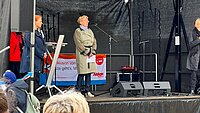 Menschen auf einer Bühne bei einer Kundgebung, im Vordergrund eine Person mit blauem Pullover mit der Aufschrift 'Teilhabe ist Menschen Recht'