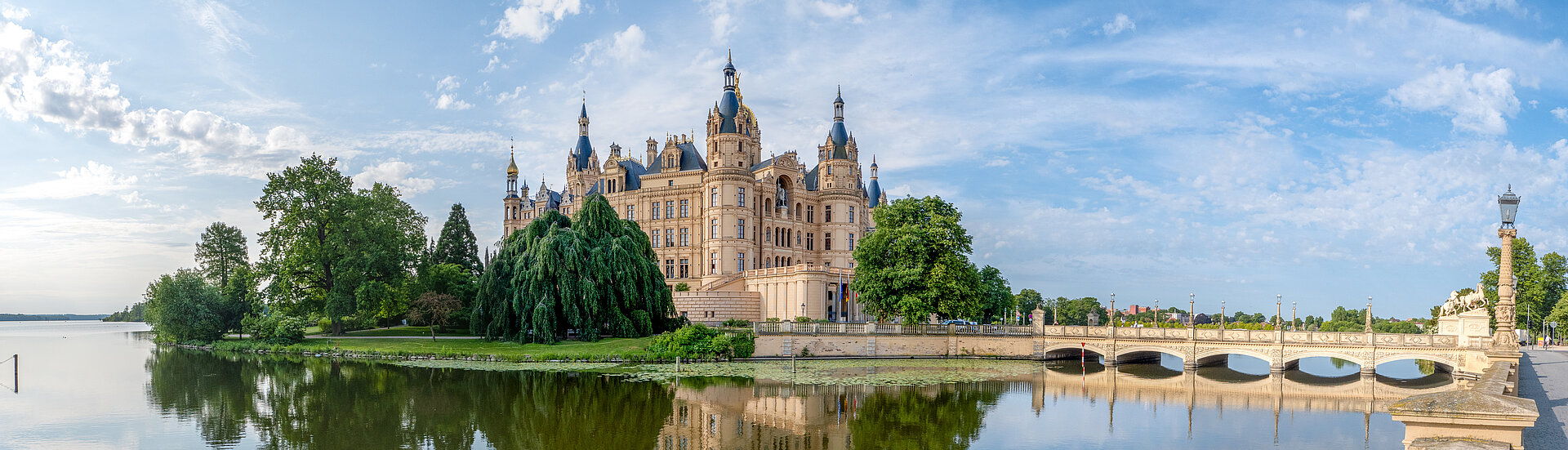 Panoramablick auf das Schweriner Schloss mit Türmen und reich verzierter Fassade, umgeben von Bäumen und Wasser, im Vordergrund eine steinerne Brücke über den Burgsee.
