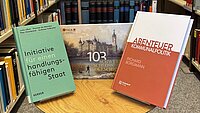 Drei neu Bücher der Landtagsbibliothek sind vor einem Bücherregal arrangiert und werden als aktuelle Neuerwerbungen präsentiert.