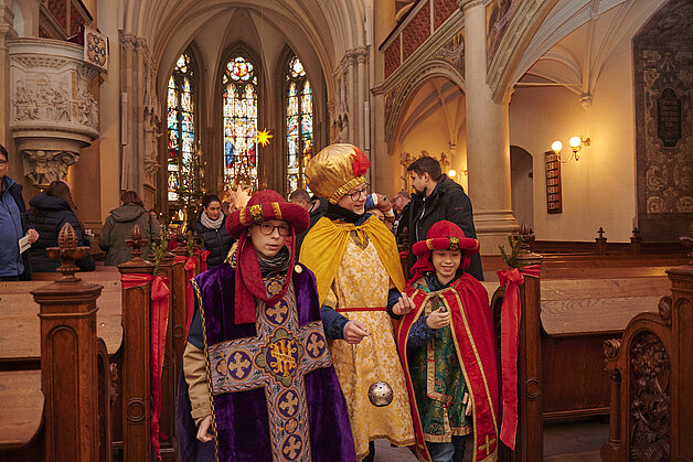 Drei als Sternsinger verkleidete Kinder in farbenfrohen Gewändern stehen in der festlich geschmückten Schweriner Schlosskirche. Im Hintergrund bunte Kirchenfenster und weitere Besucherinnen und Besucher.“