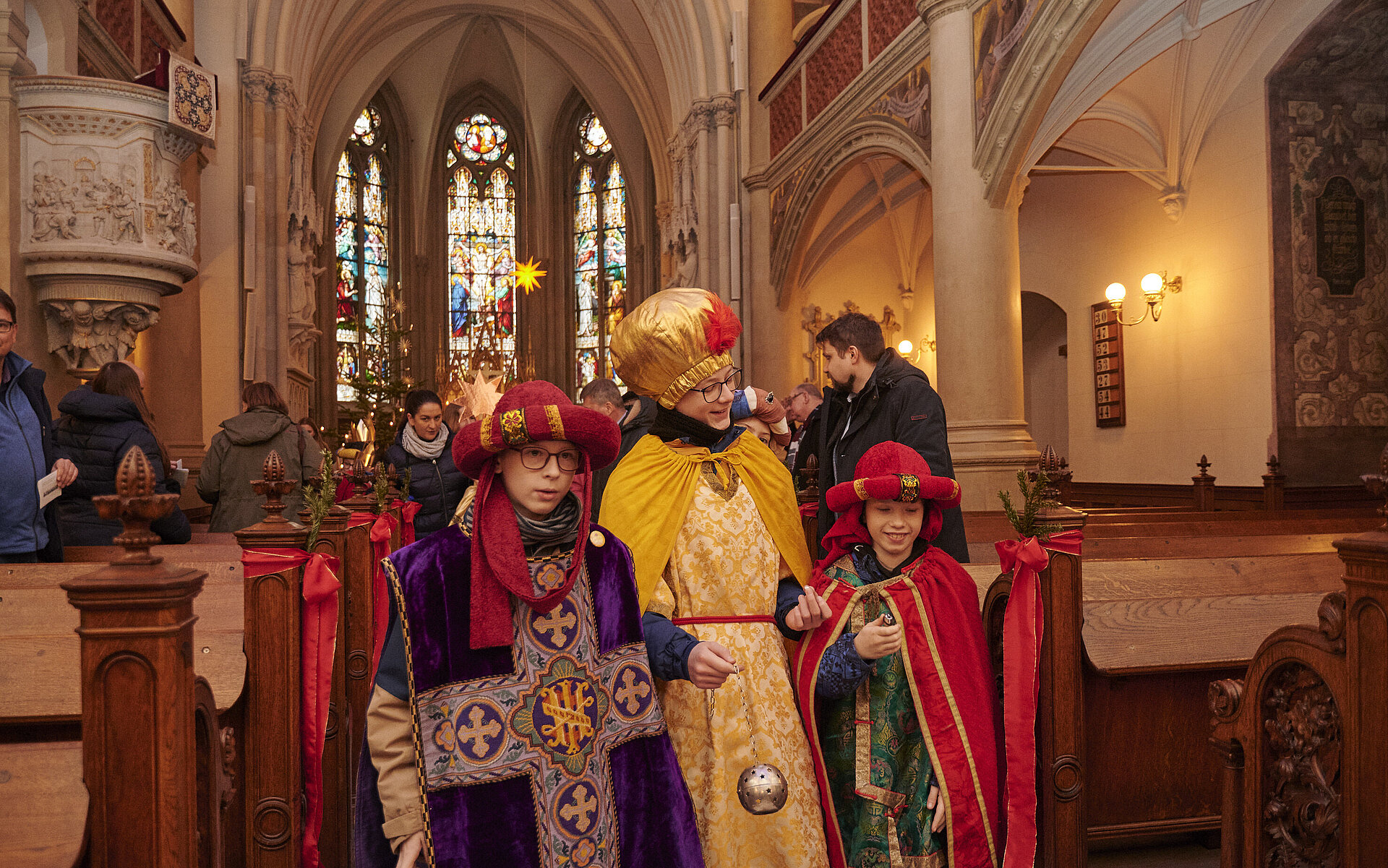 Drei als Sternsinger verkleidete Kinder in farbenfrohen Gewändern stehen in der festlich geschmückten Schweriner Schlosskirche. Im Hintergrund bunte Kirchenfenster und weitere Besucherinnen und Besucher.“