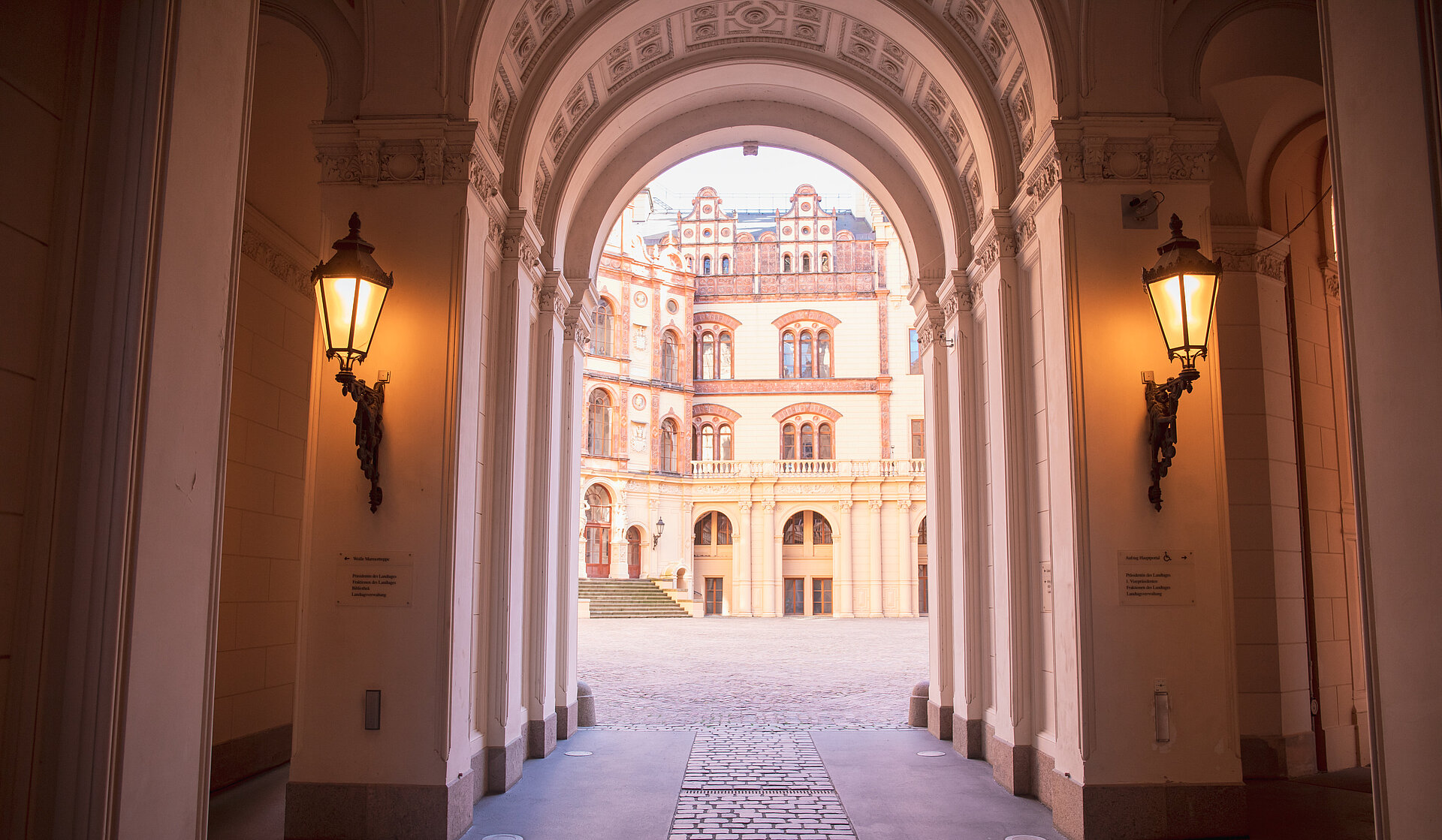Blick durch einen beleuchteten Torbogen mit kunstvoll verzierten Säulen und Decken auf den Innenhof des Schweriner Schlosses mit historischer Fassade im Hintergrund.