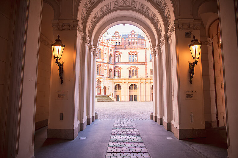 Blick durch einen beleuchteten Torbogen mit kunstvoll verzierten Säulen und Decken auf den Innenhof des Schweriner Schlosses mit historischer Fassade im Hintergrund.
