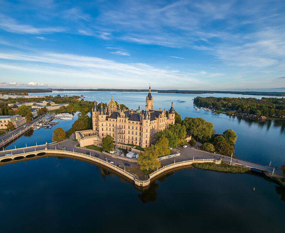 Luftaufnahme des Schweriner Schlosses, das von Wasser umgeben auf einer Insel im Schweriner See liegt. Umgeben von Bäumen, Brücken und angrenzenden historischen Gebäuden, erstreckt sich im Hintergrund die Stadt Schwerin.