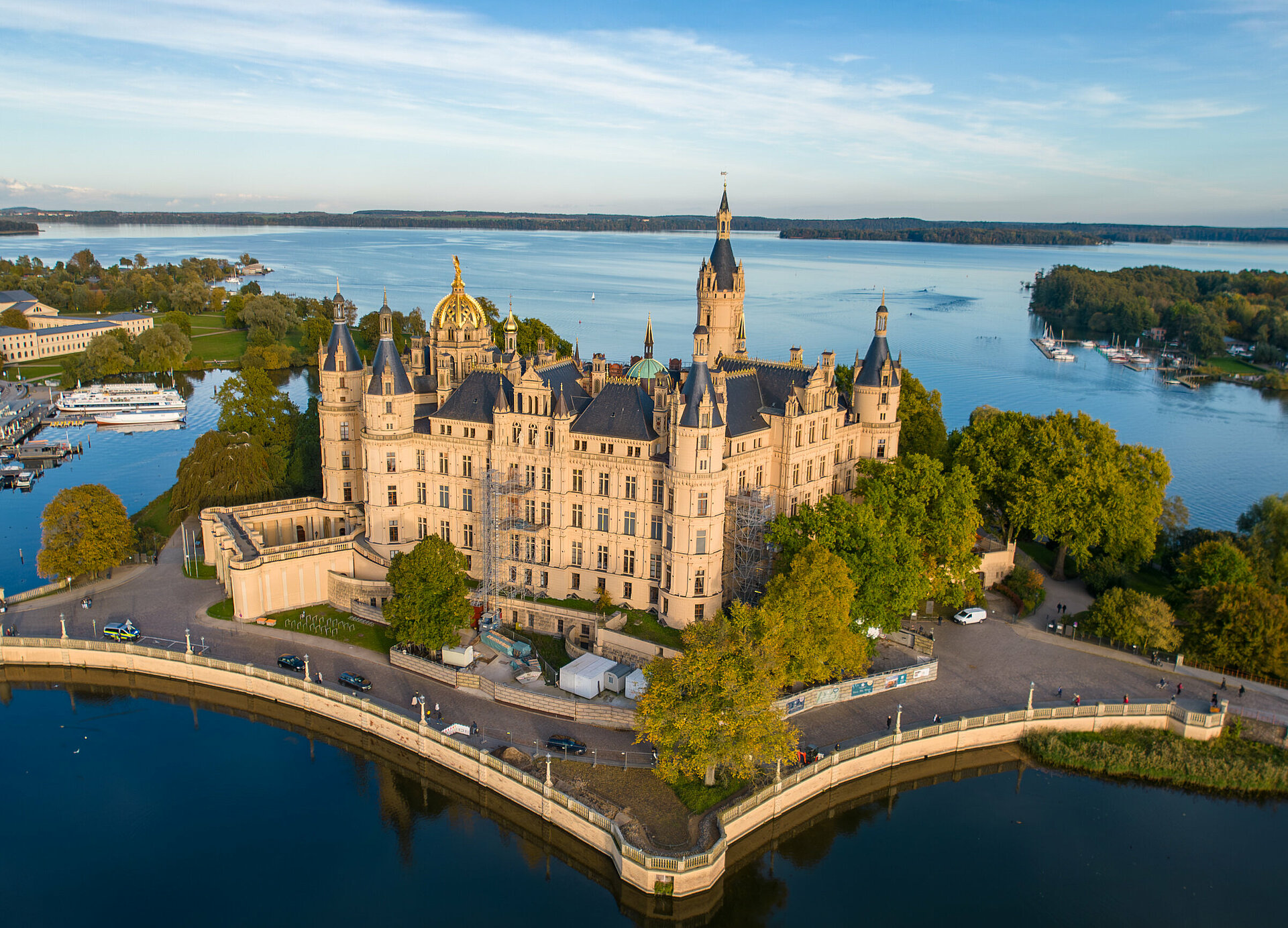 Luftaufnahme des Schweriner Schlosses, das von Wasser umgeben auf einer Insel im Schweriner See liegt. Umgeben von Bäumen, Brücken und angrenzenden historischen Gebäuden, erstreckt sich im Hintergrund die Stadt Schwerin.