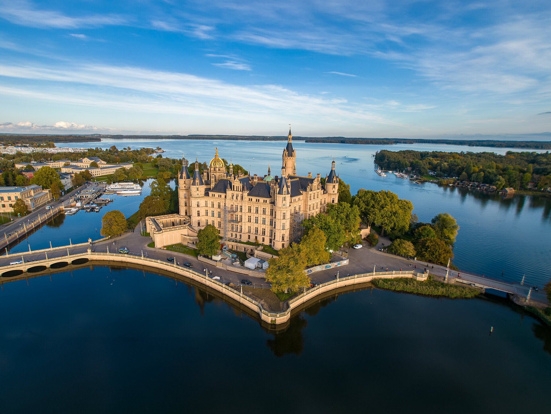 Luftaufnahme des Schweriner Schlosses, das von Wasser umgeben auf einer Insel im Schweriner See liegt. Umgeben von Bäumen, Brücken und angrenzenden historischen Gebäuden, erstreckt sich im Hintergrund die Stadt Schwerin.