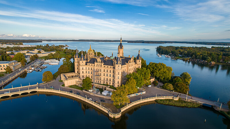 Luftaufnahme des Schweriner Schlosses, das von Wasser umgeben auf einer Insel im Schweriner See liegt. Umgeben von Bäumen, Brücken und angrenzenden historischen Gebäuden, erstreckt sich im Hintergrund die Stadt Schwerin.