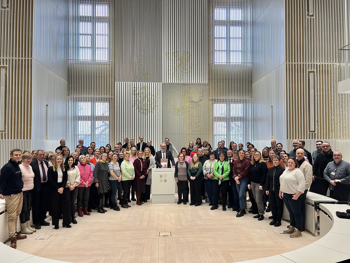 Gruppenfoto einer großen Gruppe Menschen im Plenarsaal, zu einem halben Kreis angeordnet