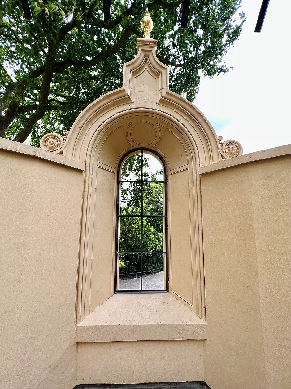 Barockes Fenster mit geschwungener Rahmung und Blick ins Grüne, eingebaut in eine Schlossmauer des Schweriner Burggartens