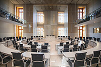 Leerer Plenarsaal des Landtags Mecklenburg-Vorpommern mit modernen, halbrund angeordneten Tischen und schwarzen Drehstühlen vor einer hellen Lamellenwand mit hohen Fenstern