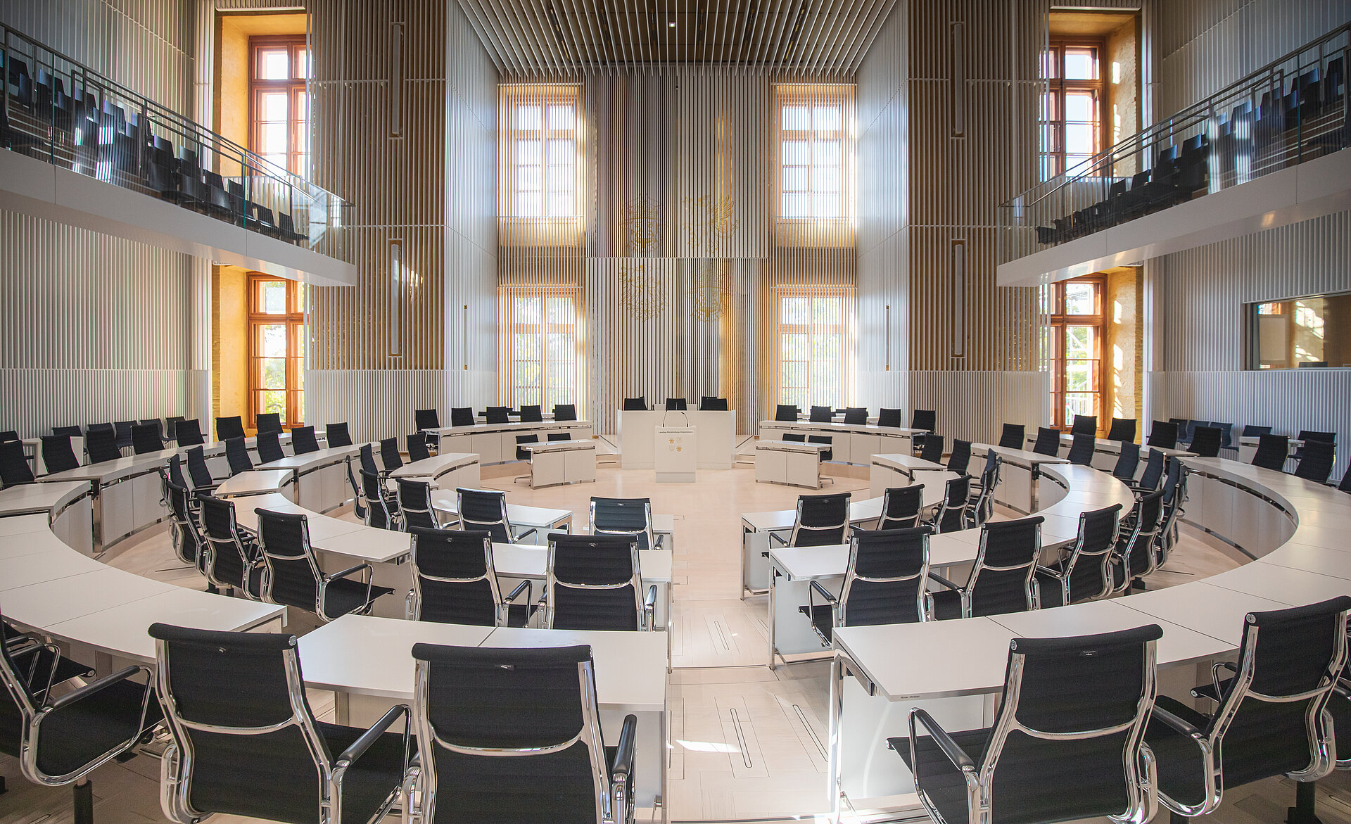 Leerer Plenarsaal des Landtags Mecklenburg-Vorpommern mit modernen, halbrund angeordneten Tischen und schwarzen Drehstühlen vor einer hellen Lamellenwand mit hohen Fenstern