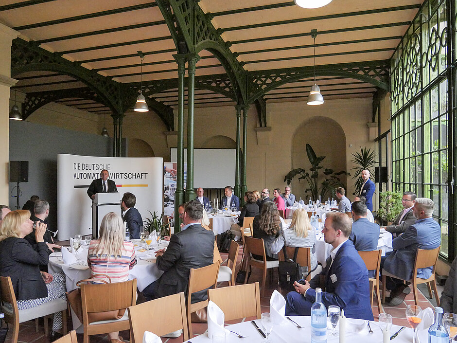 Parlamentarischer Abend in einem lichtdurchfluteten Saal mit hohen Decken und gusseisernen Säulen; Gäste an runden Tischen, während eine Rednerin spricht am Pult vor dem Banner „Die Deutsche Automatenwirtschaft“