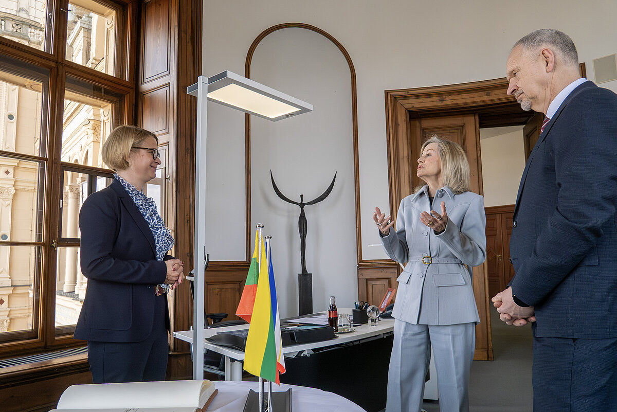 Zwei Frauen und ein Mann unterhalten sich in einem repräsentativen Raum, Flaggen stehen auf dem Tisch, eine Kunstskulptur ist im Hintergrund zu sehen