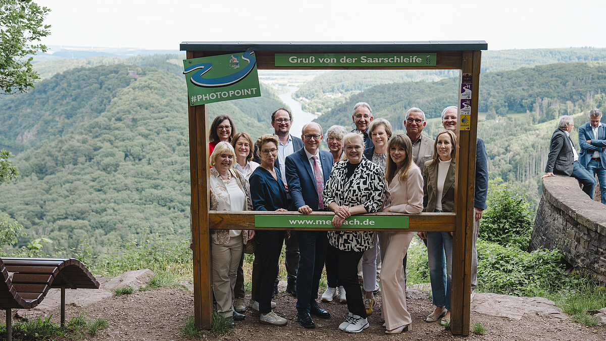 Gruppe von Menschen steht hinter einem großen Holzrahmen mit der Aufschrift 'Gruß von der Saarschleife' vor bewaldeter Flusslandschaft