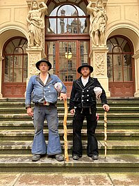 Zwei Männer in traditioneller Wanderkleidung mit Wanderstäben stehen vor einer historischen Treppe mit großen Skulpturen und einem Gebäude mit großen Fenstern