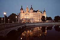 Abendliche Ansicht des beleuchteten Schweriner Schlosses mit Spiegelung im Schlossgraben und historischer Brücke im Vordergrund.