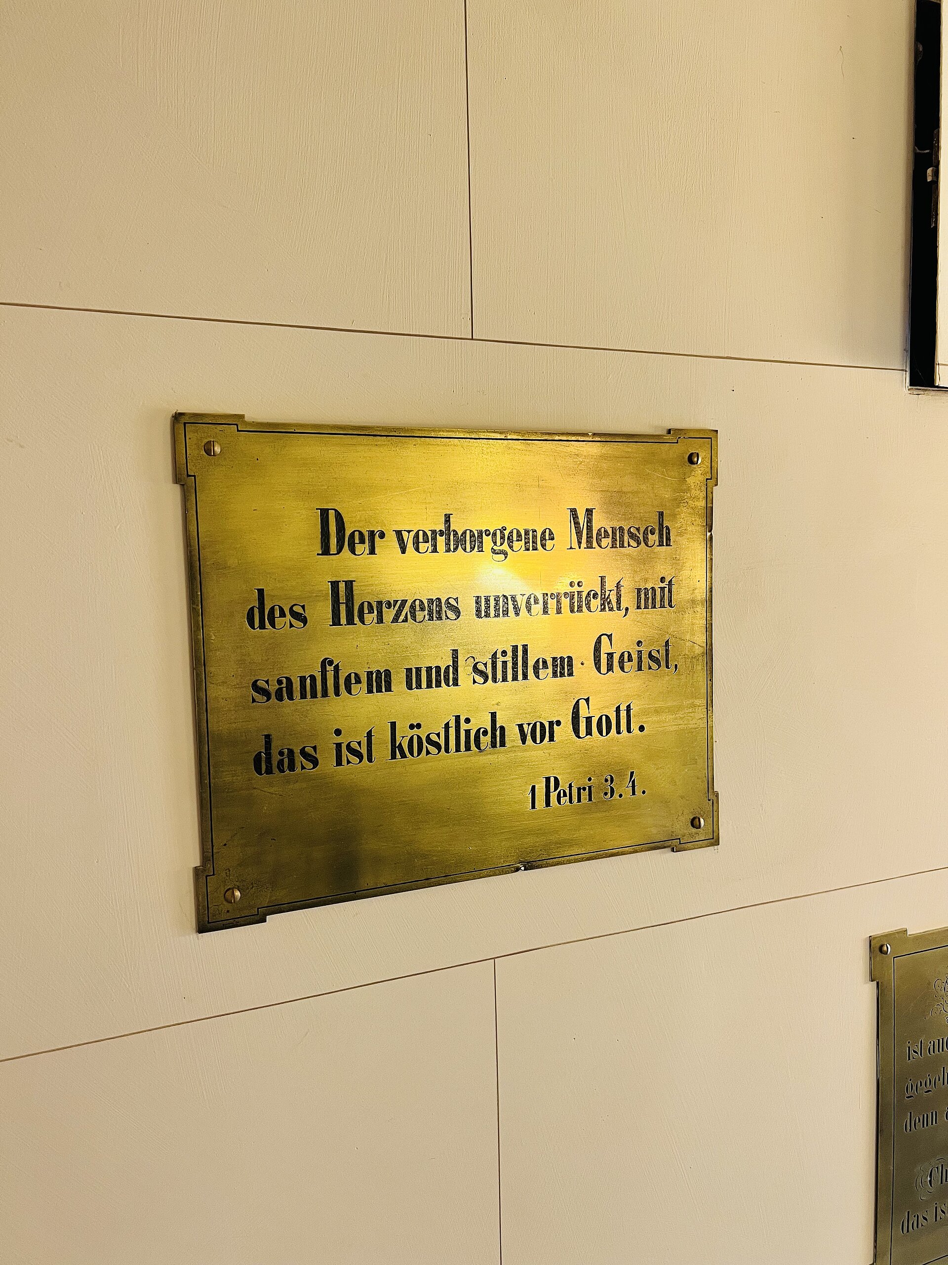 Eine goldene Gedenktafel mit der Bibelinschrift „Der verborgene Mensch des Herzens unverrückt, mit sanftem und stillem Geist, das ist köstlich vor Gott. 1. Petrus 3,4.“ an einer hellen Kirchenwand.