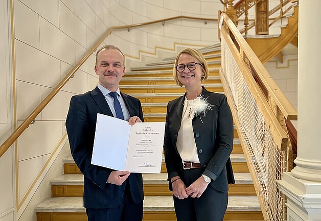 Ein Mann hält eine Urkunde in die Kamera, eine ihm von der Frau daneben überreicht wurde